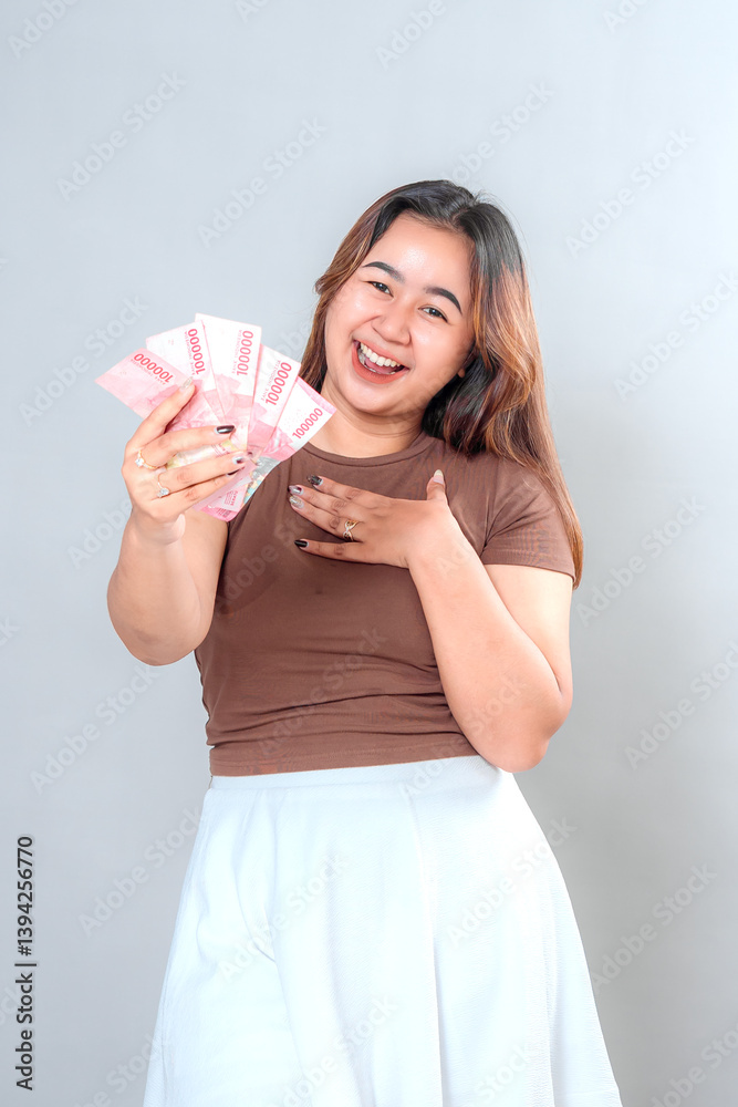 A cheerful young woman smiling brightly while holding Indonesian rupiah banknotes, expressing happiness, excitement, and financial success on a plain background. A cheerful young woman smiling brightly while holding Indonesian rupiah banknotes, expressing happiness, excitement, and financial success on a plain background.