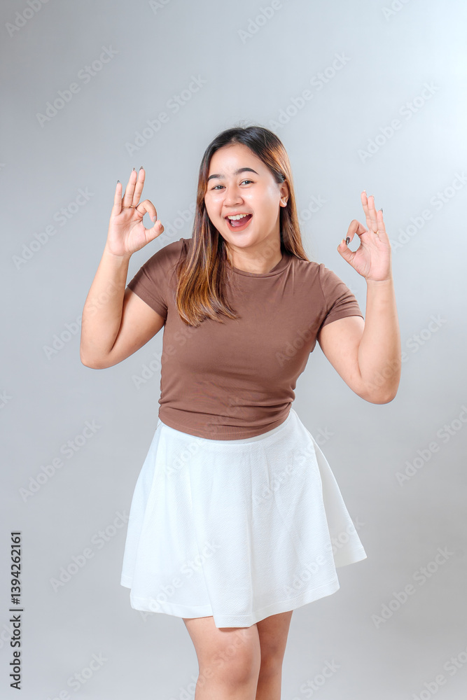 Cheerful young woman smiling brightly while showing double OK hand gesture. Her joyful facial expression conveys confidence, agreement, and positive communication for advertising use. Cheerful young woman smiling brightly while showing double OK hand gesture. Her joyful facial expression conveys confidence, agreement, and positive communication for advertising use.
