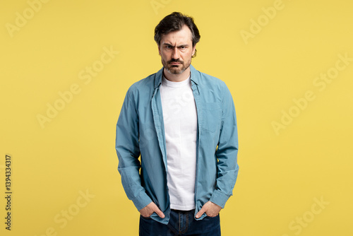 Grumpy man frowning and holding hands in pockets on yellow background