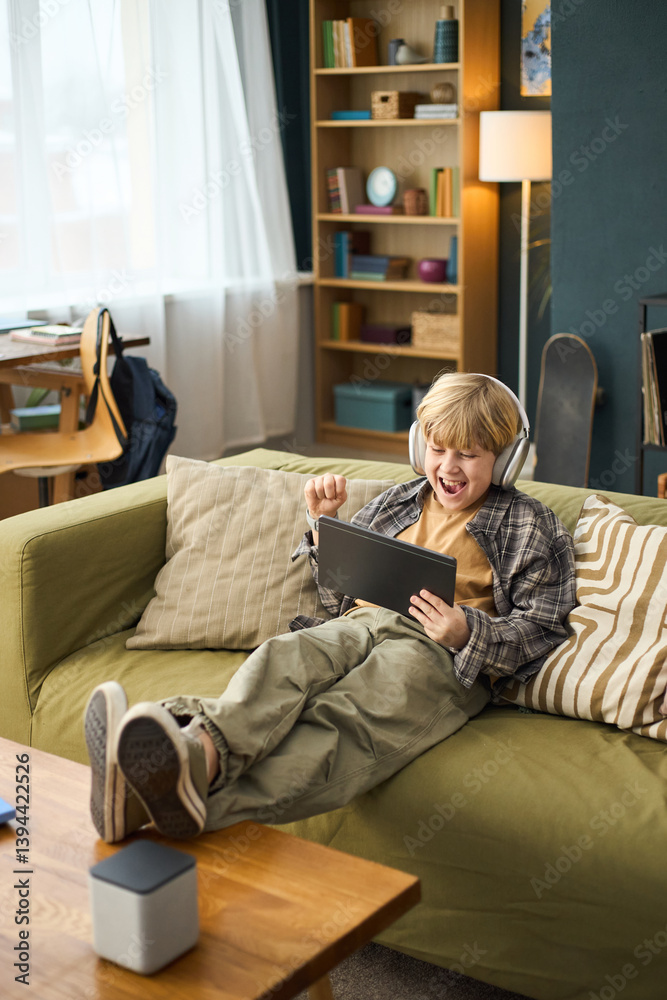 Obraz premium Young boy sitting on couch wearing headphones and using tablet, displaying joyful expression, relaxing in cozy living room with shelves and lamp