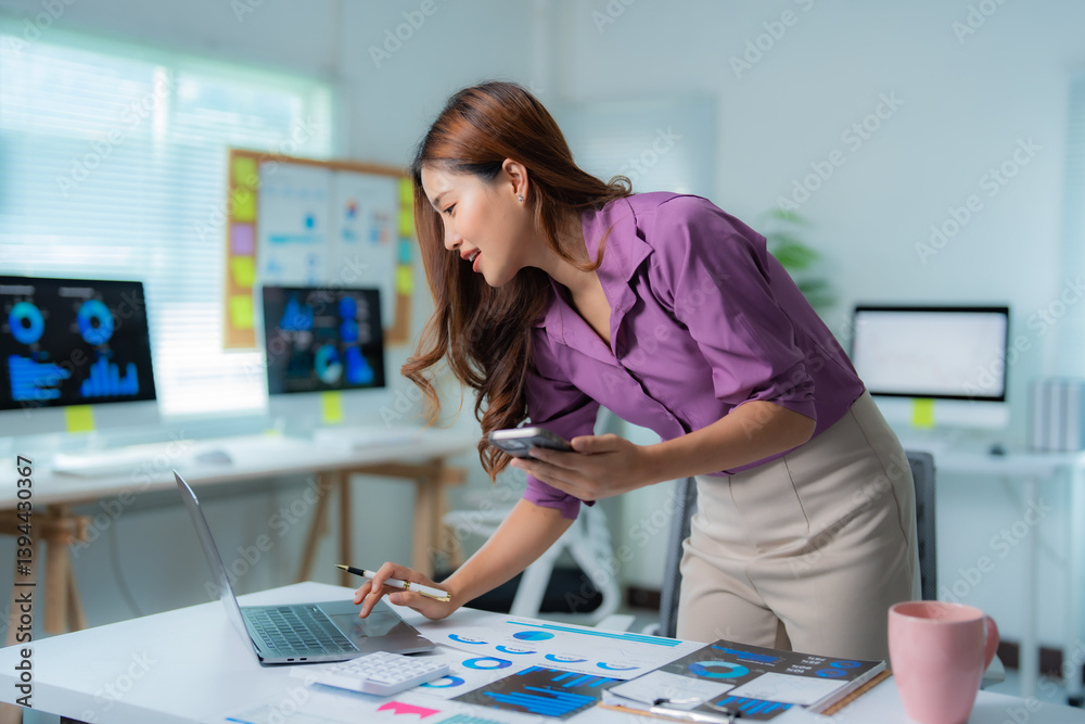Fototapeta premium Young businesswoman standing at her desk analyzing financial charts displayed on laptop and smartphone, working with graphs and data in a modern office environment