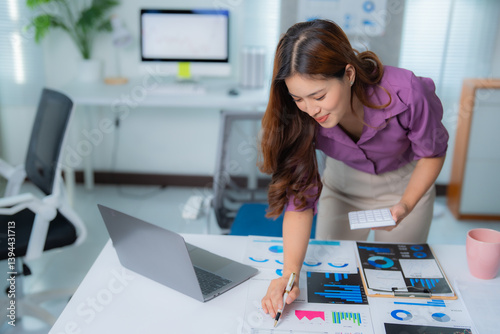 Young asian businesswoman holding calculator in hand and pointing at charts while analyzing marketing data in modern office with laptop and desktop computer