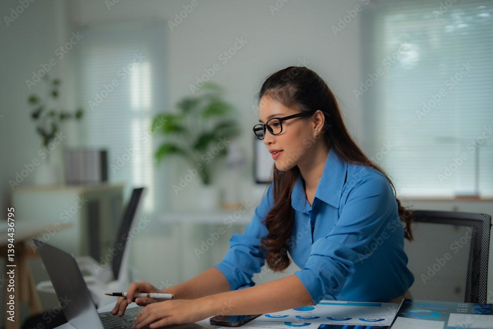 Obraz premium Young Asian businesswoman wearing glasses and blue shirt working with laptop and analyzing financial charts at office desk, holding pen in her right hand