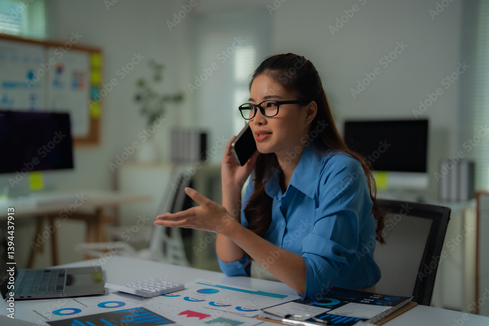 Fototapeta premium Young professional woman wearing glasses and blue shirt is sitting at desk in office, talking on phone and discussing charts, using laptop and calculator