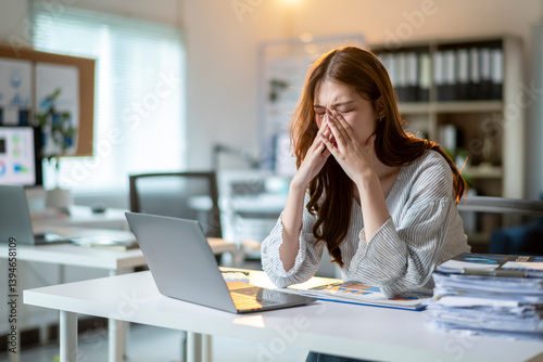 A woman is sitting at a desk with a laptop and a stack of papers