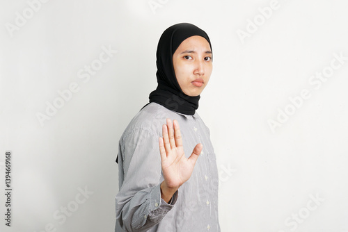 Young Asian woman raising her hand with palm facing forward in a stop gesture, expressing refusal or warning with a serious expression while standing against a plain background.