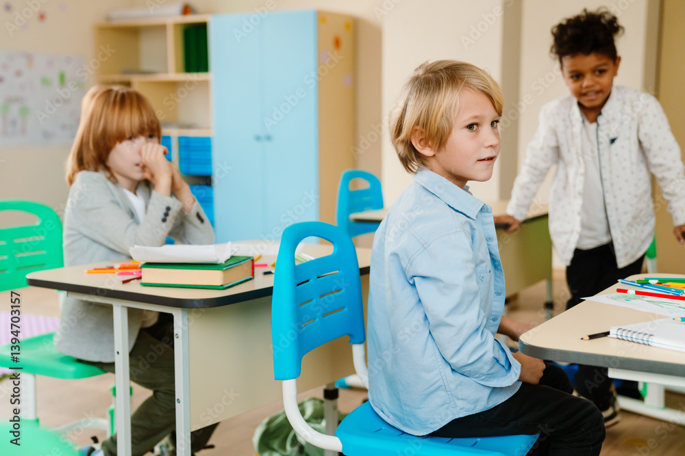 Obraz premium A White school-age boy sits at his desk, his White female classmate sits behind him, and their Black classmate stands beside them during a school break in a classroom. Concept of a school friendship.