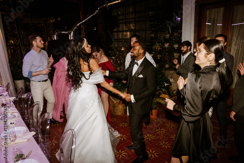 Obraz na płótnie A Black young bride and groom dancing together on a red tiled floor surrounded by a multinational group of wedding guests celebrating during a lively indoor wedding reception with plants