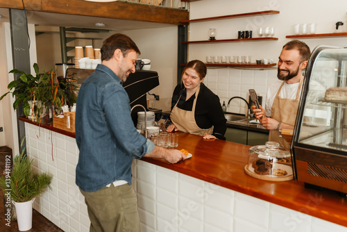 A white adult man receives food at a coffee shop counter from two smiling cafe workers, a woman and a man wearing aprons. They are engaged in a friendly conversation.