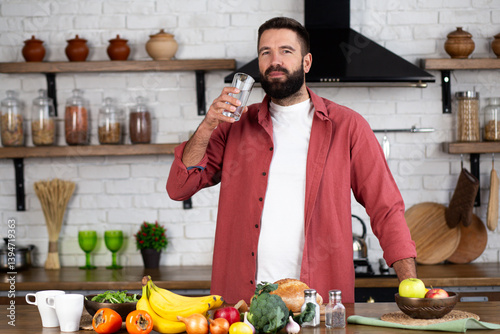 A young man stands in kitchen and drinks water from a glass
