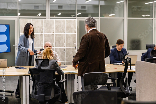 A middle-aged white-skinned businessman, standing near his seat at a long table where his colleagues gather, in an office room, talking to his mid-30s Asian female colleague, who stands across