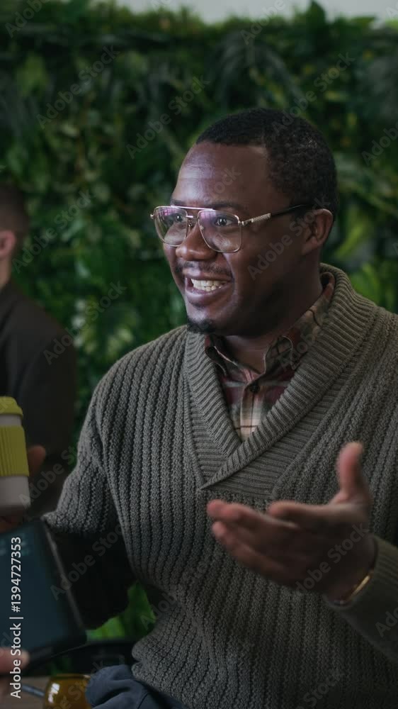 Vertical of young inspired Black man talking to unrecognizable colleague at modern green office space during coffee break Vertical of young inspired Black man talking to unrecognizable colleague at modern green office space during coffee break