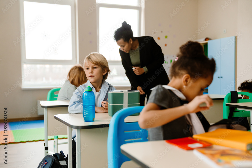 A diverse group of students, including a White boy and a Black girl, interacts with an adult Latino female teacher in a classroom. The room includes colorful furniture and supplies for students. A diverse group of students, including a White boy and a Black girl, interacts with an adult Latino female teacher in a classroom. The room includes colorful furniture and supplies for students.