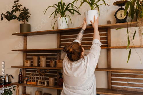A view from the back of an overweight white-skinned woman in her late 20s, with tattoos on her hand, standing on her toes and putting a plant onto the top shelf of a cozy caf? interior