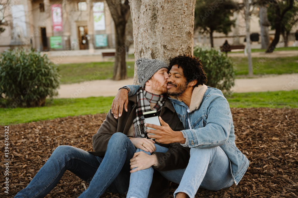 Fototapeta premium An African American man aged 35, wearing a denim jacket, sitting on a field in a park with his mid-30s White husband, and taking a selfie with his phone as the husband kisses him on the cheek