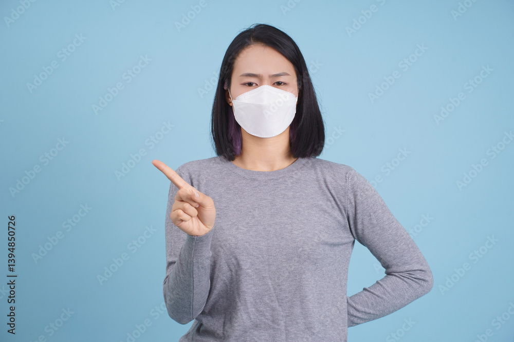 Woman in gray long-sleeve shirt pointing while wearing a mask against a solid blue background during indoor setting Woman in gray long-sleeve shirt pointing while wearing a mask against a solid blue background during indoor setting