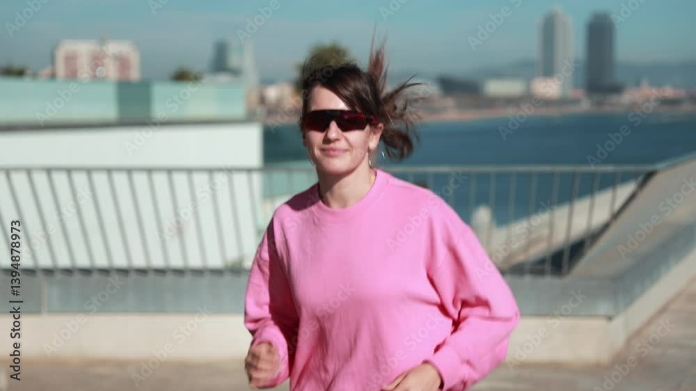 A White sportswoman in her 20s wearing a pink sweatshirt and sunglasses, is jogging outdoors, smiling as her hair blows in the wind, with a scenic urban backdrop of palm trees and the ocean. A White sportswoman in her 20s wearing a pink sweatshirt and sunglasses, is jogging outdoors, smiling as her hair blows in the wind, with a scenic urban backdrop of palm trees and the ocean.