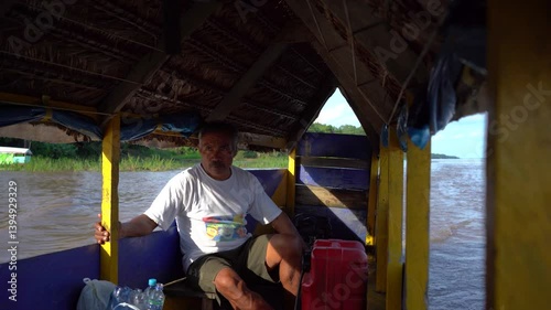 Driver of a long wooden boat with a roof steering the motor in the Amazon River near Iquitos, Peru