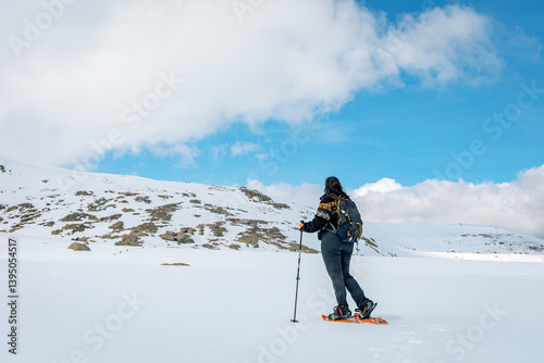 Female snowshoer hiking toward a snow-covered ridge in Sierra de Gredos, Spain, with trekking poles and backpack under a bright blue sky with puffy white clouds in a remote alpine setting