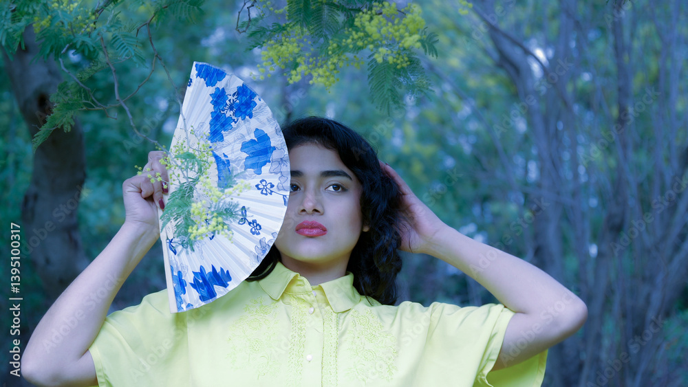 Model posing with a hand fan partially covering her face, standing in a natural setting with mimosa branches
