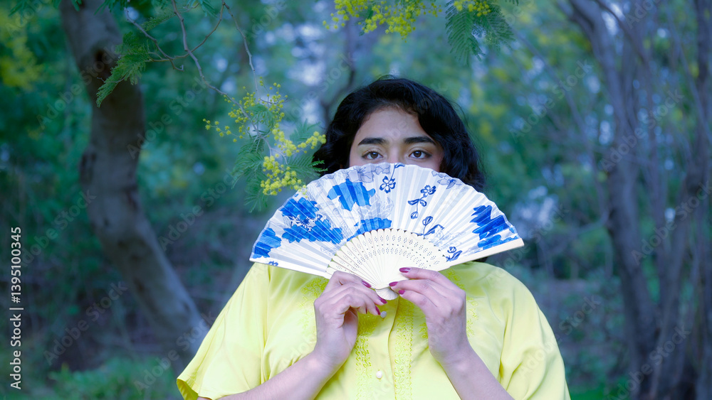 Woman holding a decorated hand fan in front of her face in a park with mimosa tree