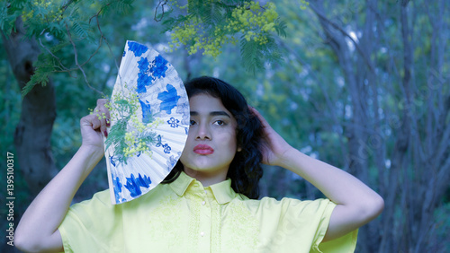 Obraz na płótnie Model posing with a hand fan partially covering her face, standing in a natural setting with mimosa branches w salonie