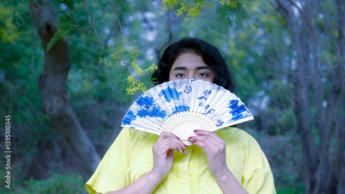 Obraz na płótnie Woman holding a decorated hand fan in front of her face in a park with mimosa tree w salonie