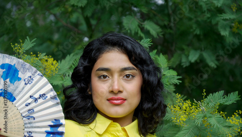 Beautiful young hispanic woman holding a hand fan, posing in a green lush garden