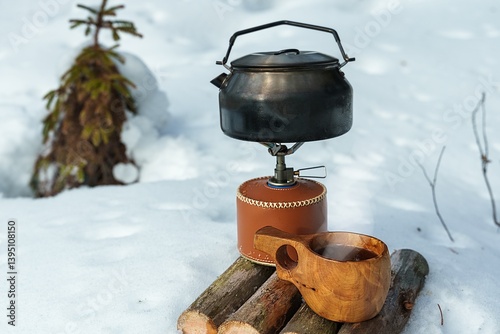 A camping kettle sits on a portable gas stove. Hot coffee in winter. A traditional wooden kuksa cup.
