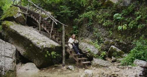 Russia, Caucasus. A beautiful young woman of European appearance uses the phone while sitting on the steps of a wooden staircase in the mountains among the rocks overgrown with vegetation, near stream