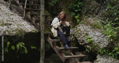 Russia, Caucasus. A beautiful young woman of European appearance uses the phone while sitting on the steps of a wooden staircase in the mountains among the rocks overgrown with vegetation, near stream