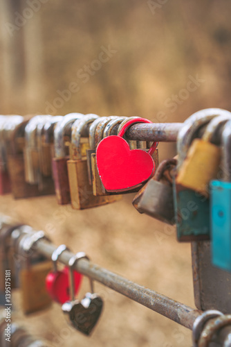A red heart-shaped love padlock placed among many others on a metal fence. Blurred background.