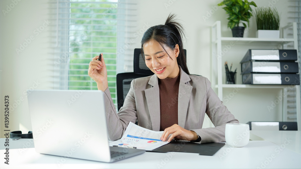 Happy young professional woman looks at printed charts while holding a pen at her bright, modern desk. Happy young professional woman looks at printed charts while holding a pen at her bright, modern desk.