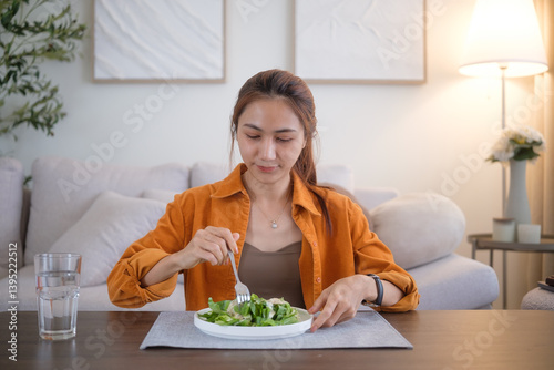 Asian woman in an orange shirt eating salad with a glass of water nearby, captured in a warm, inviting home environment.