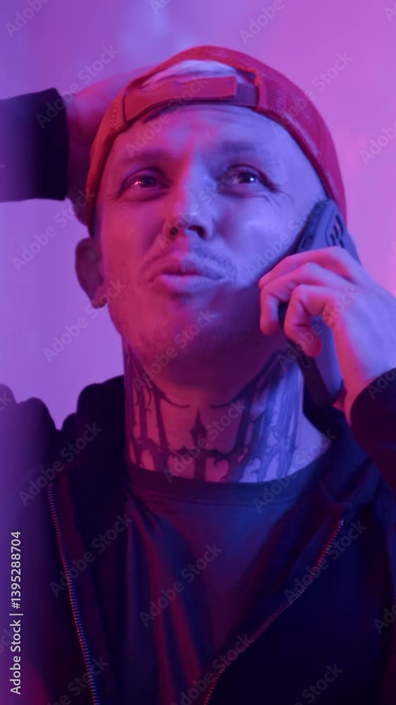 Vertical neon shot of male tattooed cybercrime talking on mobile phone in low key room