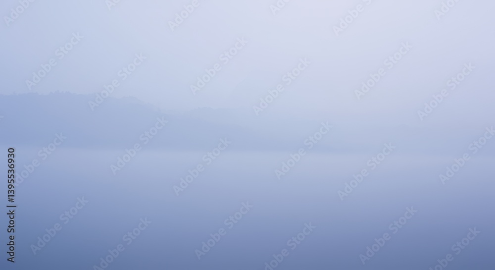 Fototapeta premium Serene fog-covered lake at dawn with distant mountains and soft reflections
