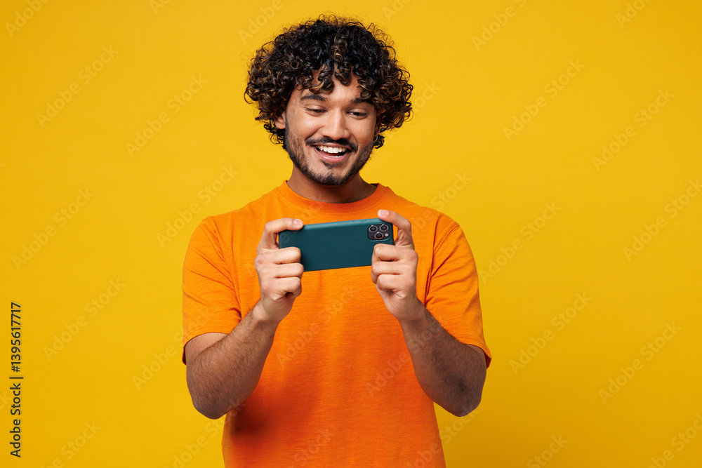 Young gambling Indian man wear orange t-shirt casual clothes use play racing app on mobile cell phone hold gadget smartphone for pc video games isolated on plain yellow background. Lifestyle concept. Young gambling Indian man wear orange t-shirt casual clothes use play racing app on mobile cell phone hold gadget smartphone for pc video games isolated on plain yellow background. Lifestyle concept.