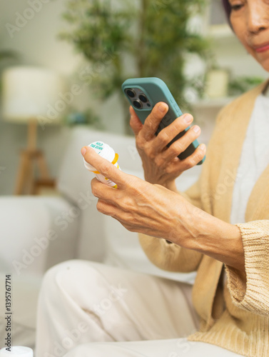 Asian senior hand holding bottle of pill checking prescription using mobile phone searching prescription and information online on internet, online video call with doctor in living room at home.