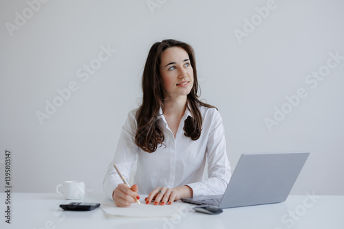 Professional woman writing notes while working on a laptop in a minimalist office setting during daylight hours
