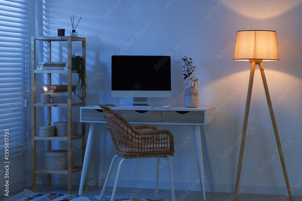Comfortable workplace with computer on desk and floor lamp at home office in evening