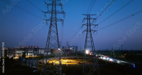 Aerial view of high voltage electricity transmission towers and power lines standing over an illuminated highway interchange against the night sky.