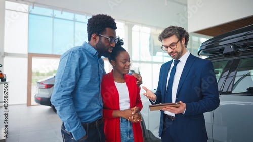 Handsome auto dealer helping mixed-raced customers to choose car that fits their needs and budget. Man in business costume holding tablet device and talking with married couple in vehicle centre.