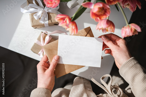 Female hands holding blank white card above kraft envelope with wrapped gifts and pink tulips arranged on white table. Greeting card mockup