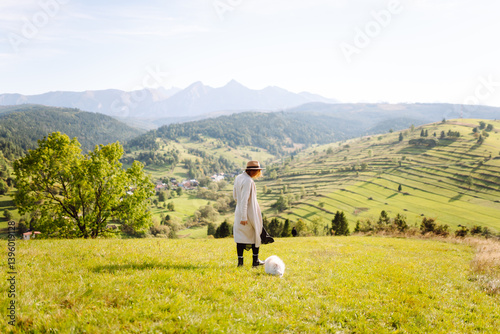 Obraz na płótnie Female tourist having fun with her small white dog on grassy hillside. Back view of beautiful woman with her pet enjoying mountain landscape at sunset. Freedom, travel and fun concept. w salonie