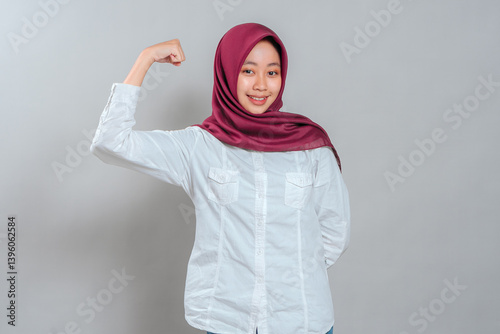 Confident young woman in maroon hijab smiling while flexing her arm muscle, showing strength and empowerment. Her happy and proud expression conveys positivity and determination.