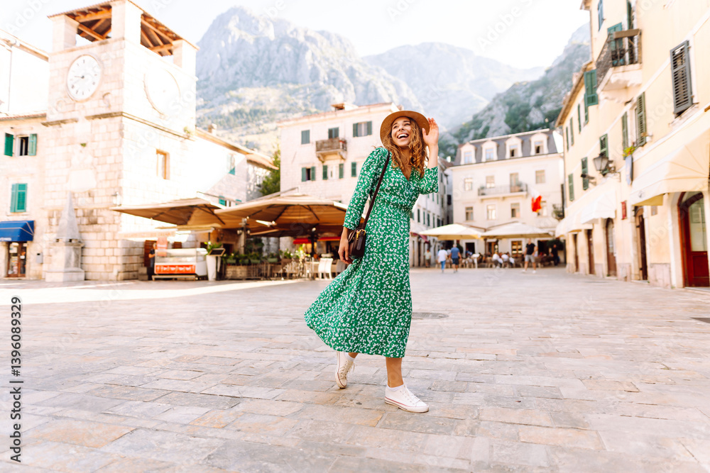 Naklejka premium A beautiful woman in a bright dress and hat walks through the old streets of Europe on a sunny day. A young tourist enjoys the sunrise outdoors. Tourism concept. Active lifestyle.