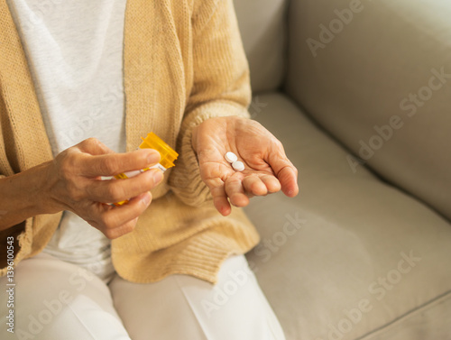 Asian Senior Woman hand with pill in hand pouring medicine from medication bottle sitting on sofa in living room at home. Senior woman with medicine, taking pills, vitamin for treatment herself.