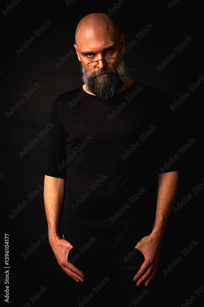 Intense bald man with a grey beard and round glasses making a small pinch gesture. Low key portrait with dramatic lighting and expressive face on black background