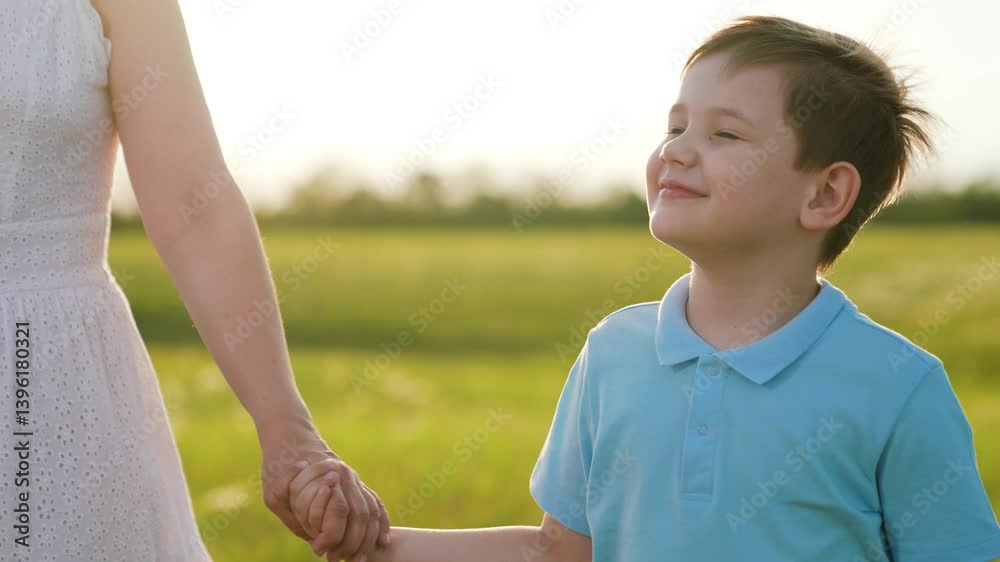 Mother son walking together in nature park. Boy smiling holding mother's hand, walk with mother through sunny summer green field close-up. Happy family. Mother son child holding hands enjoying nature Mother son walking together in nature park. Boy smiling holding mother's hand, walk with mother through sunny summer green field close-up. Happy family. Mother son child holding hands enjoying nature