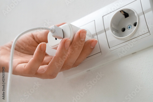 Female hand unplugging charger from socket on light wall, closeup photo